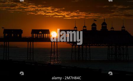 Die goldene Kugel der aufgehenden Sonne erscheint zwischen zwei Gebäuden auf dem silhouettierten Eastbourne Pier bei Sonnenaufgang mit unterbeleuchteten Wolken im Hintergrund Stockfoto