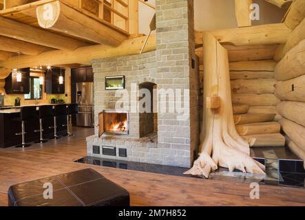 Brauner Lederottoman und beleuchteter Kamin aus grauem Backstein, Treppe aus weißem Kiefernholz und Baumstamm mit sichtbaren Wurzeln. Stockfoto