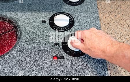 Temperaturregler mit männlicher Hand auf der Kochfläche des Ofens. Warnleuchte für die Sicherheit der Küche bei heißer Oberfläche am Gerät auf der Kochplatte. Stockfoto