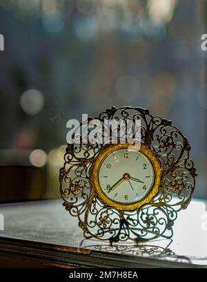 Bokeh-Hintergrund zeigt die Tischuhr im Retro-Stil auf einem weißen Tisch im Vordergrund an. Die Uhr besteht aus altem Goldmetall mit dekorativem Fadenkreuz auf einem Drahtständer. Stockfoto