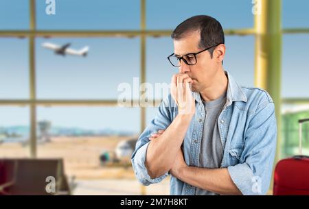 Erwachsener lateinamerikanischer Mann mit Brille am Flughafen, der gestresst und nervös aussah, mit der Hand auf dem Mund, der sich die Nägel beißt, und Angststörung wegen Angst vor Stockfoto