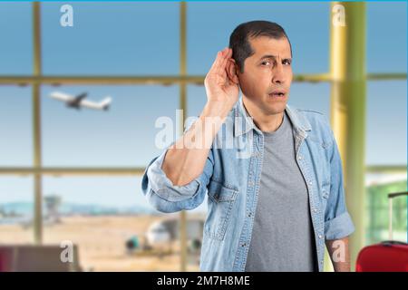 Studiofoto eines gutaussehenden Mannes, der am Flughafen lächelt und Hand über Ohr hört und Gerüchte oder Klatsch hört. Schwerhörigkeitsbegriff. Stockfoto