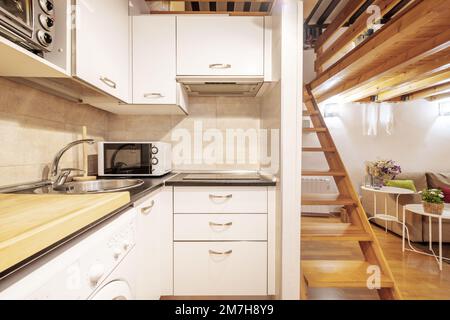 Kleine offene Küche mit weißen Schränken, weiße Geräte in einem Loft-Apartment mit Treppe und Kiefernkassettendecke Stockfoto