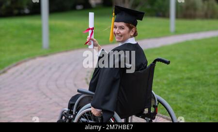 Glückliche weiße Frau im Rollstuhl dreht sich um und hält ihr Diplom draußen. Stockfoto