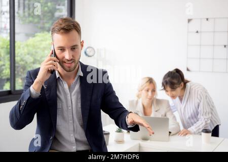 Porträt eines jungen Geschäftsmanns, der mit seinem Smartphone lächelt und spricht, während Mitarbeiter oder Kollegen im Büro, Manager oder Manager mit arbeiten Stockfoto