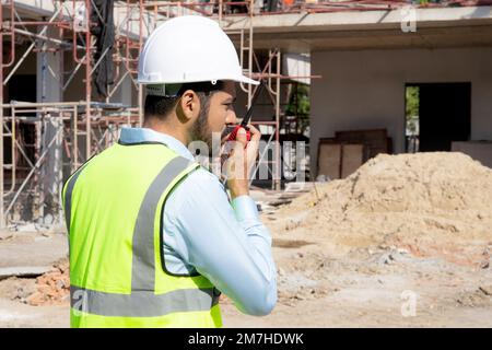 Ein junger Ingenieur, der Funkbefehle mit einem Arbeiter auf der Baustelle, einem Architekten oder einem Auftragnehmer verwendet, spricht mit dem Funkgerät, um die Entwicklung von Steuerungs- und Planungsfunktionen zu steuern Stockfoto