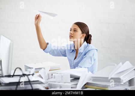 Der Tag ist einfach nicht schnell genug. Eine junge Geschäftsfrau, die ein Papierflugzeug warf, während sie an ihrem Schreibtisch saß. Stockfoto