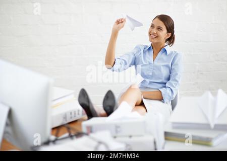 Etwas abgelenkt an ihrem Schreibtisch. Eine junge Geschäftsfrau, die ein Papierflugzeug warf, während sie an ihrem Schreibtisch saß. Stockfoto