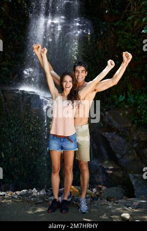 Wir lieben unseren geheimen Ort im Dschungel. Porträt eines wunderschönen jungen Paares, das vor einem Wasserfall in einem Wald jubelt. Stockfoto