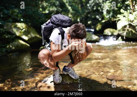 Anhalten für eine erfrischende Pause. Ein junger Mann spritzt kühles Wasser auf sein Gesicht, während er in der Nähe eines Bachs wandert. Stockfoto