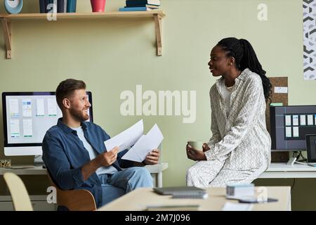 Seitliches Porträt zweier junger Kollegen, die im Büro über das Projekt sprechen und lächeln Stockfoto