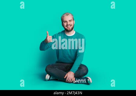 Großes Foto eines jungen Mannes, der auf dem Boden sitzt, zeigt Daumen-hoch-Bewilligung isoliert auf blauem Hintergrund. Positive Person. Stockfoto