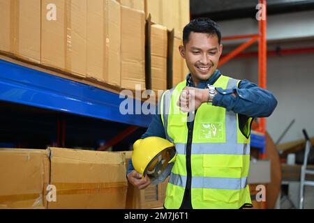 Lächelnder asiatischer männlicher Vorgesetzter, der die Zeit auf seiner Uhr überprüft, während er im Lagerhaus steht Stockfoto