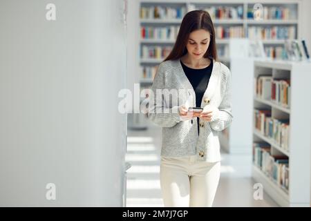 Telefon, Bibliothek und Frau vernetzen sich über soziale Medien, mobile Apps oder das Internet mit Mobilgeräten. Bücher, Wissen und junge Studentinnen, die schreiben oder Stockfoto