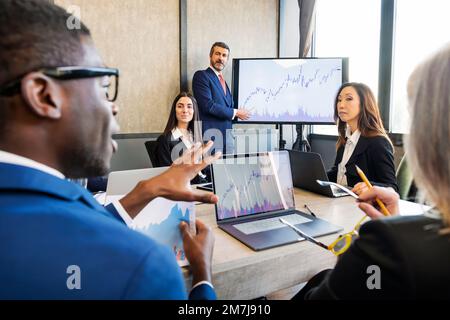 Gruppe multiethnischer Kollegen, die sich mit einem Laptop an einem Tisch versammeln, während sie während der Präsentation über ein Finanzprojekt sprechen Stockfoto