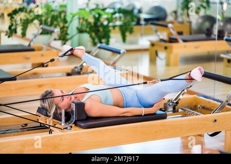 Ganzkörperfrauen in Sportbekleidung liegen auf dem Reformerbett und machen geteilte Übungen mit Riemen während des Pilates Trainings im Light Studio Stockfoto