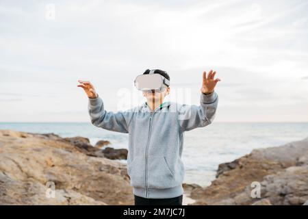Ein Junge trägt einen Virtual-Reality-Headset-Simulator auf einem Felsbrocken am Meer Stockfoto