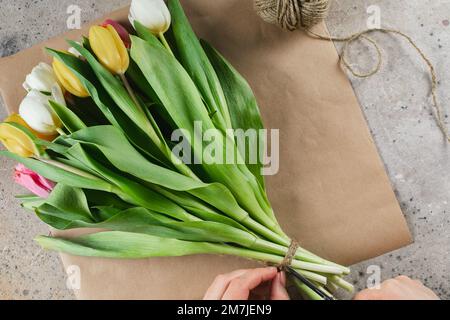 Weiße und gelbe Tulpen in einem Paket Handwerk. Weibliche Hände arrangieren einen Blumenstrauß in Handwerkspapier. Frühjahrsferien-Konzept. Stockfoto