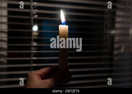 Brennende Kerze in den Händen vor dem Hintergrund eines Fensters mit Jalousien (Nahaufnahme). Stromausfall. Energiekrise. Stromausfallkonzept Stockfoto