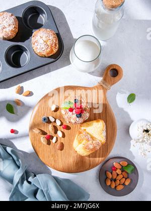 Frisch gebackene hausgemachte Muffins mit Nüssen und Beeren auf einem Holzbrett auf einem hellen Tisch mit Milch und Mandeln. Das Konzept eines nahrhaften und gesunden Stockfoto