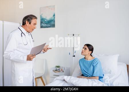Senior Doktor mit Papiermappe, die mit brünetten Patienten auf der Krankenhausstation spricht, Stock Image Stockfoto