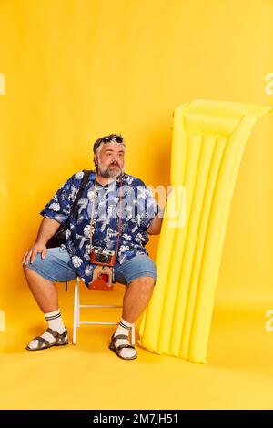 Porträt eines fetten, reifen Mannes im stilvollen Hemd, der auf einer Schwimmmatratze sitzt und sich auf einem hellgelben Studiohintergrund posiert Stockfoto
