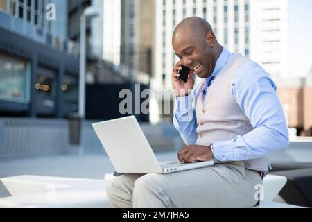 Eleganter afroamerikanischer Geschäftsmann, der im Freien mit einem Laptop telefoniert. Stockfoto