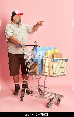 Porträt eines erwachsenen Mannes in legerer Kleidung, der Einkaufen geht, mit einem Einkaufswagen posiert und einen pinken Studiohintergrund hat. Verkauf Stockfoto