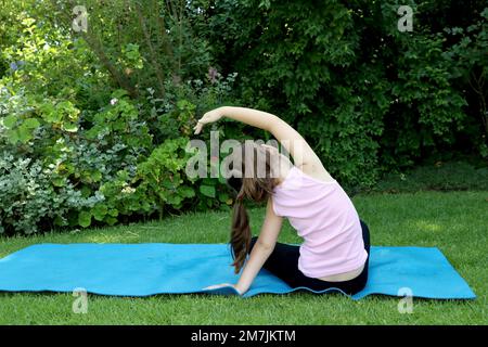Ein 7-10-jähriges Mädchen, das Yoga macht, posiert im Garten und trägt Zöpfe auf einer blauen Yogamatte Stockfoto