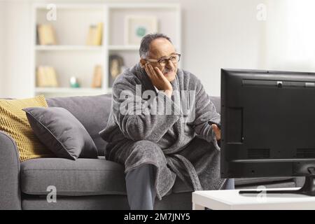 Gelangweilter, alter Mann im Bademantel, sitzt auf dem Sofa, hält eine fernbedienung und sieht zu Hause fern Stockfoto