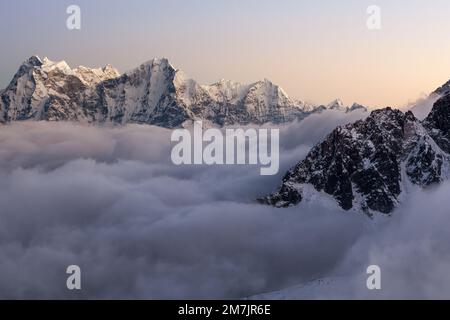 Malerische Berglandschaft des Himalaya. Dramatische schneebedeckte Gipfel, die sich über dem Bergtal erheben und bei Sonnenuntergang mit lockigen Wolken gefüllt sind. Stockfoto