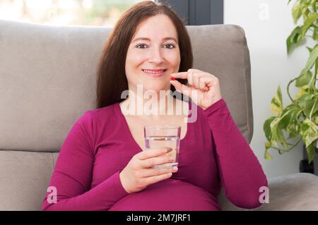 Glückliche schwangere Frau, die zu Hause eine rote Pille nimmt Stockfoto