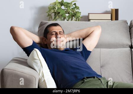 Glücklicher Mann, der sich mit Armen auf dem Kopf entspannt und in der Nacht zu Hause auf einer Couch sitzt Stockfoto