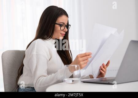 Lächelnde junge Geschäftsfrau mit Brille, die sich die Dokumente ansieht und mit dem Notizbuch nachsieht. Stockfoto
