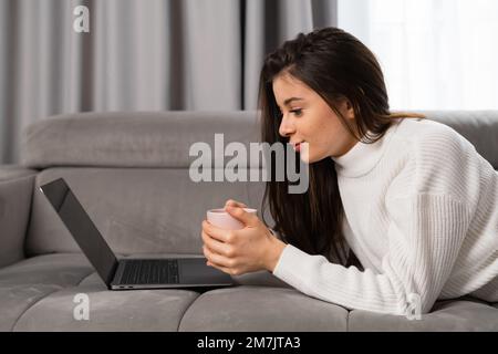 Junge, schöne Frau, die sich entspannt, eine Tasse hält, sich einen Film mit einem Laptop ansieht, auf dem Sofa liegt Stockfoto