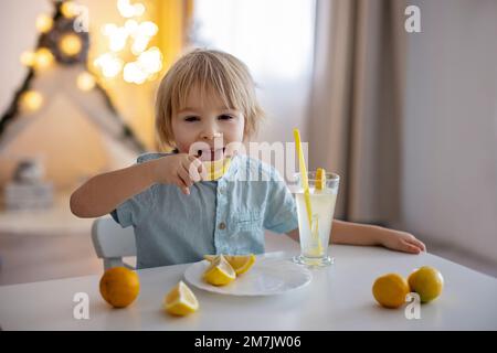 Niedliches Kleinkind, blonder Junge, lecken Zitronen zu Hause und trinken Limonade, lustige Gesichter machen Stockfoto