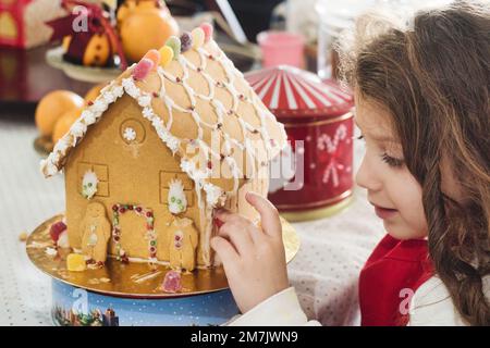 Ein junges weißes Mädchen, das einem hausgemachten Lebkuchenhaus den letzten Schliff gibt Stockfoto