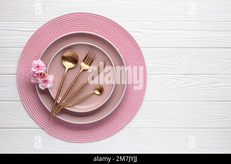 Rosafarbenes Geschirr und goldenes Besteck mit Blumen, Stille auf weißem Holztisch. Draufsicht. Geschirr und Utensilien zum Servieren und Essen. Moderner Tiger Stockfoto