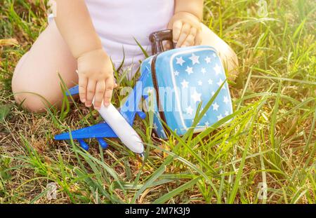 Koffer Schweinebank Flugzeug Plastikspielzeug auf gelben Blumen grünes Gras oder graue Decke, Kinderhände. Reisekonzept Stockfoto