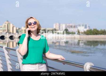 Ein lächelndes Mädchen mit Sonnenbrille, das mit Freunden telefoniert, Gesten macht, ein Date am Flussufer der Stadt macht. Das Mädchen läuft am Flussufer der Stadt entlang Stockfoto