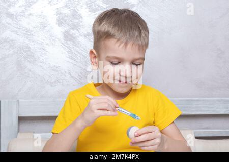 Ein kleiner blonder Junge malt Eier für die Osterferien zu Hause. Das Kind hat Spaß und feiert den Urlaub. DIY Ostereier Konzept. Stockfoto