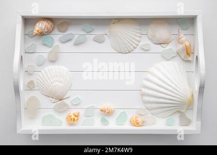 Flache Sicht auf weiße Holzschale mit Muscheln und Seeglas, Kopierraum in der Mitte. Hintergrund zu Reisezielen oder Kosmetikprodukten. Stockfoto