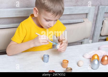 Ein kleiner blonder Junge malt Eier für die Osterferien zu Hause. Das Kind hat Spaß und feiert den Urlaub. DIY Ostereier Konzept. Stockfoto