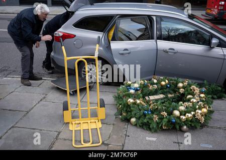 Nach den Feiertagen zu Weihnachten und Neujahr versuchen zwei Bauunternehmer, am 9. Januar 2023 in London, England, in der City of London, dem Finanzviertel der Hauptstadt, einen großen Firmenkranz in ein kleines Auto zu laden. Stockfoto