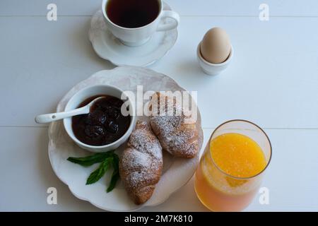 Frisches Croissant auf weißem Teller, Präsentation mit Erdbeermarmelade, Orangensaft, Ei und Tee, Draufsicht. Stockfoto