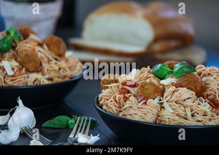 Selektive Fokussierung von zwei Spaghetti- und Fleischklößchen-Pasta mit frischen Basilikumblättern und Parmesankäse auf einem schwarzen rustikalen Holztisch. Stockfoto