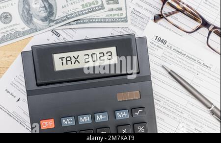 Taschenrechner mit 2023-Steuertext auf dem Bildschirm, 1040-Formular, Dollarscheine und Brille auf dem Schreibtisch. Draufsicht Stockfoto