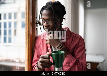 Der rücksichtsvolle Schwarze blickt aus dem Fenster, berührt das Kinn, hält den Becher fest denkt über wichtige Arbeitsaufgaben nach. Stockfoto