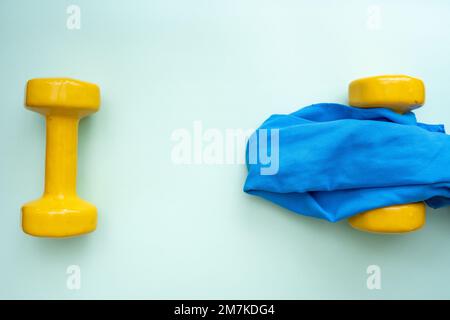 Trendige gelbe Kurzhanteln und ein kleines Handtuch mit grünem Hintergrund. Flacher Kopierbereich. Gewichtsmanagement und gesundes Lifestyle-Sportkonzept Stockfoto