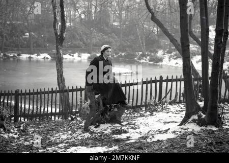 1955, historisch, im Winter und in einem schneebedeckten Wald, eine hübsche junge Frau, die einen langen Cordmantel, Hut, Handschuhe und kleine Stiefel trägt, auf einem alten Baumstumpf für ein Foto sitzt, mit einem gefrorenen See hinter ihr, Farningham, Kent, England, Großbritannien. Stockfoto